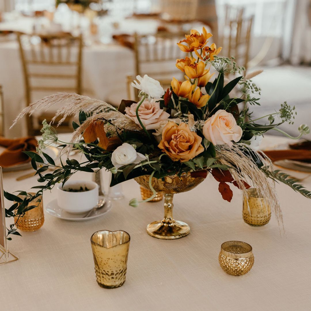 Elegant wedding centerpiece with peach roses, greenery, and dried elements in a gold vase, surrounded by candles on a table.