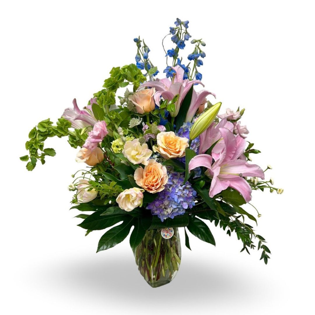 Floral arrangement featuring pastel roses, pink lilies, and blue flowers with lush greenery in a vase.