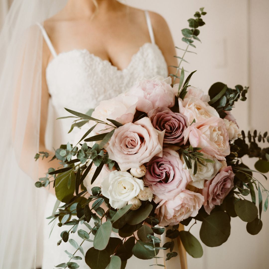 Close-up of a bride in a lace dress holding a bouquet with pastel roses and greenery.