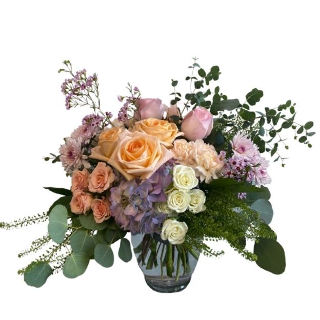 Soft pastel floral arrangement with roses, hydrangeas, and greenery in a clear glass vase.