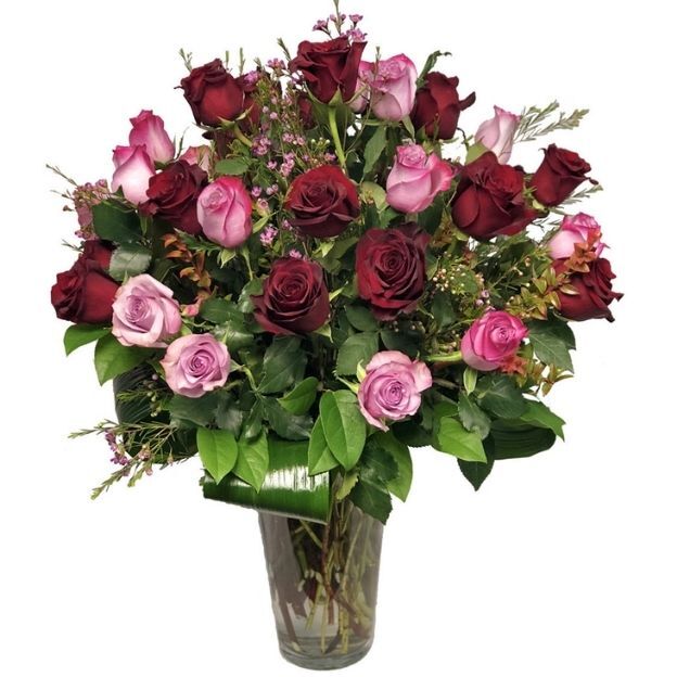 Dozens of burgundy and blush roses with leafy greens arranged in a tall cylinder vase.