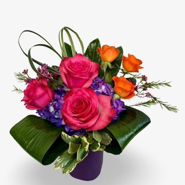 Colorful floral arrangement featuring pink and orange roses with lush greenery and purple blooms in a modern vase.