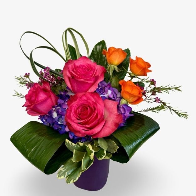 Vibrant floral arrangement with pink roses, orange roses, purple hydrangeas, and greenery in a purple vase.