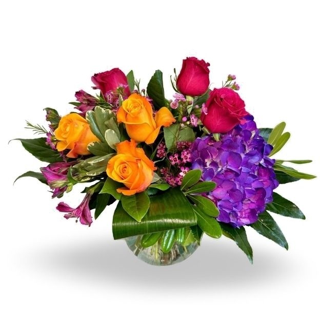 Orange and hot pink roses with purple hydrangea, pink accent flowers, and greenery in a round glass vase.