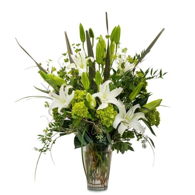 Tall bouquet of white lilies, green blooms, and long grasses with abundant greenery in a fluted clear vase.